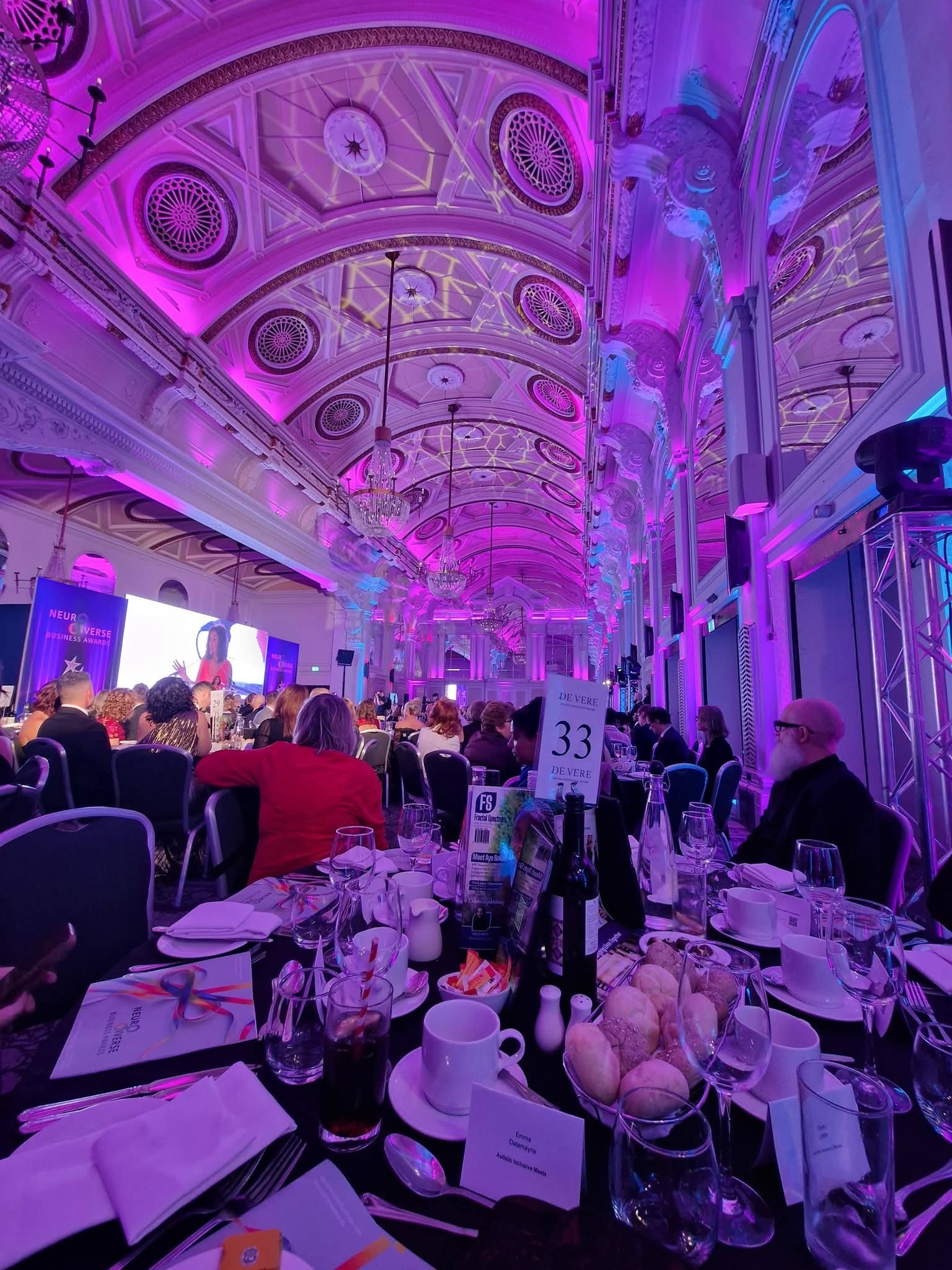 neurodiversity business venue, round table with plates, food and people sat around the tables looking ahead at the speaker on stage